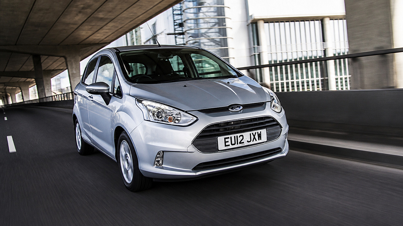 Carro prata modelo Ford em movimento em estrada urbana sob viaduto com placa EU12 JXW.