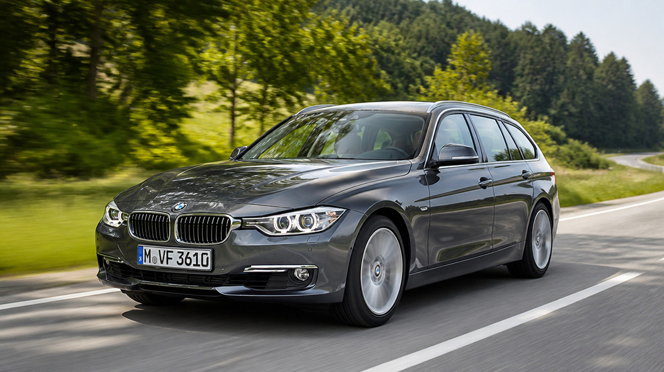 Carro BMW cinza escuro em movimento em estrada cercada por árvores verdes e campo.