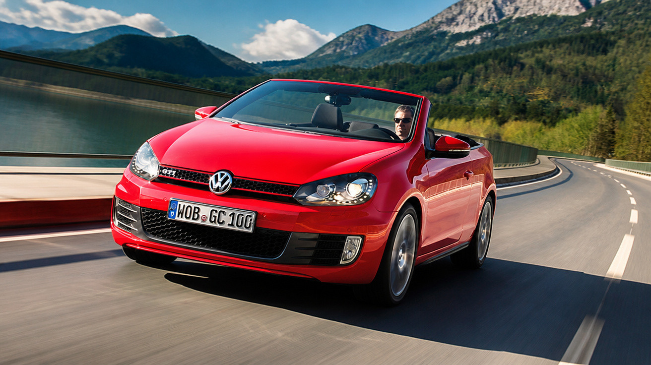 Carro conversível vermelho Volkswagen dirigido por homem em estrada sinuosa próximo a montanhas e lago.