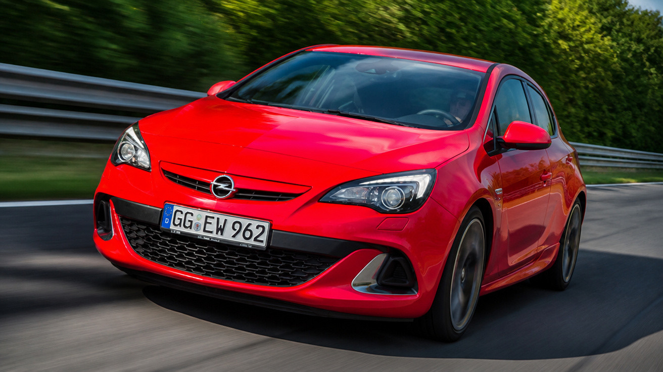 Carro esportivo vermelho em movimento em estrada com vegetação ao fundo.