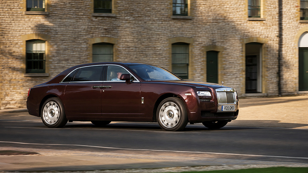 Carro de luxo Rolls-Royce marrom escuro circulando em rua com prédio de pedra ao fundo.