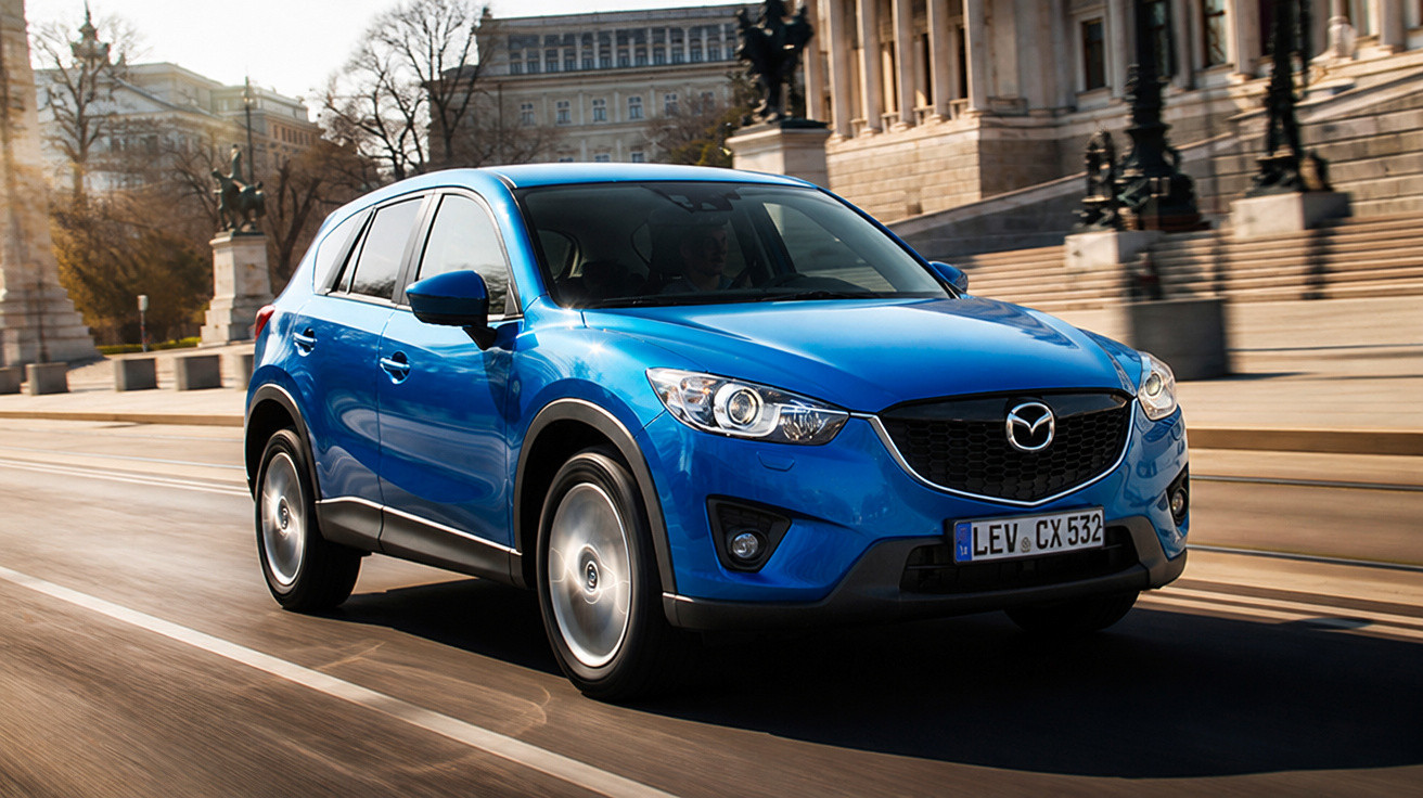 Carro SUV azul Mazda CX-5 em movimento em uma rua com edifícios históricos ao fundo.