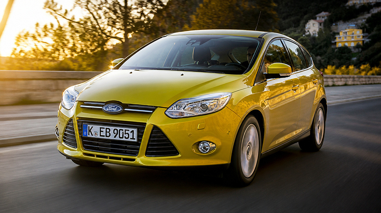 Carro Ford Focus amarelo em movimento numa estrada com paisagem ao fundo durante o pôr do sol.