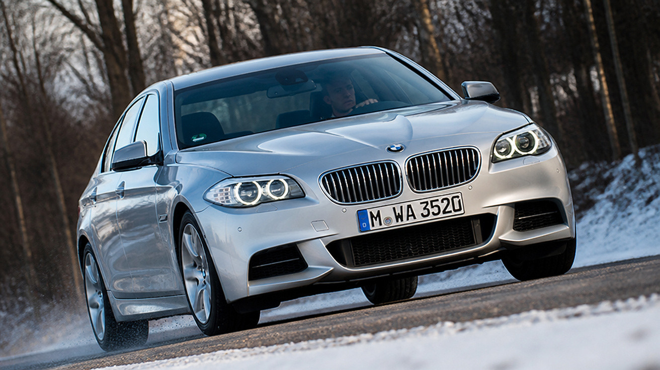 Carro sedan BMW prata em estrada com neve, em movimento próximo a uma floresta no inverno.