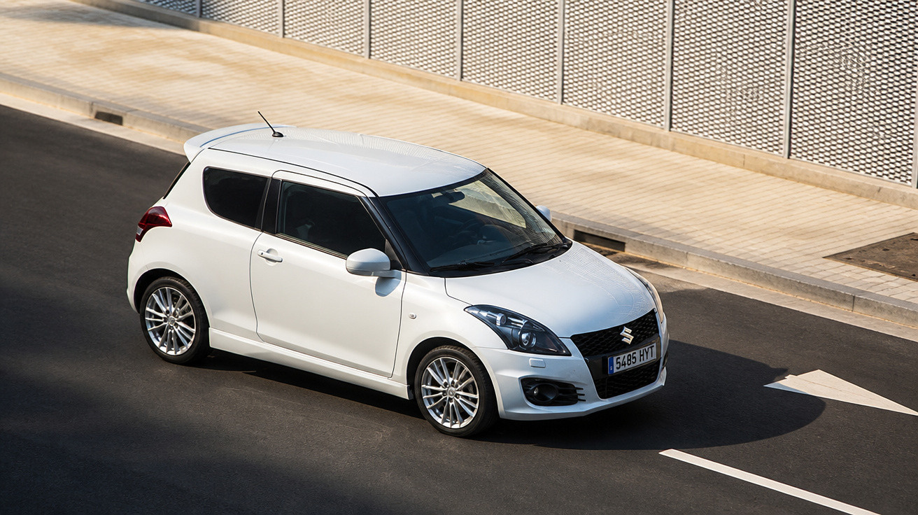 Carro branco modelo hatchback Suzuki Swift trafegando em rua asfaltada ao lado de calçada com muro de metal.