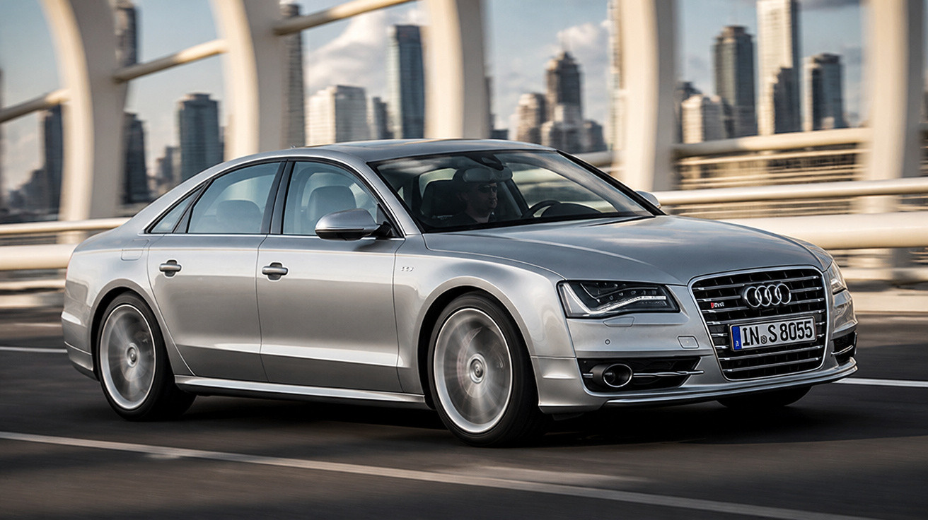Carro Audi cinza prata em alta velocidade cruzando ponte com prédios modernos ao fundo.
