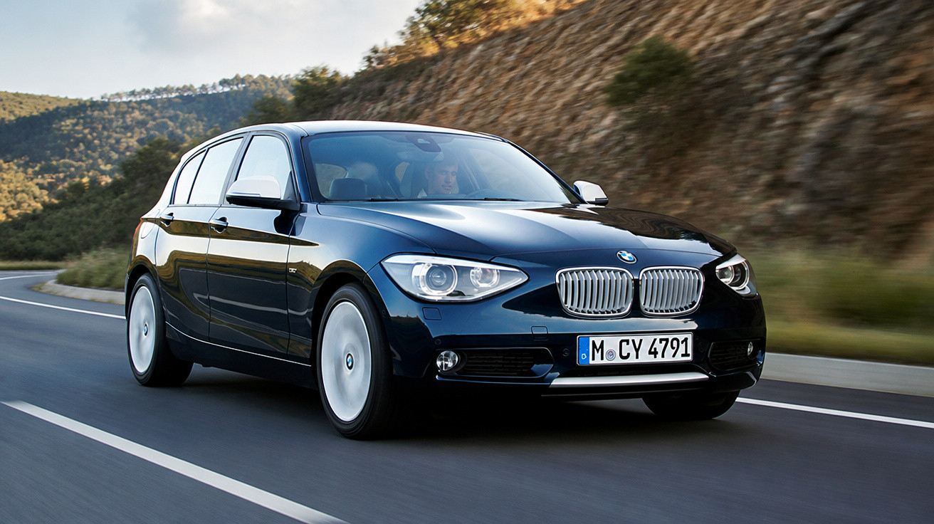 Carro BMW preto em estrada curva com paisagem montanhosa ao fundo em dia claro.