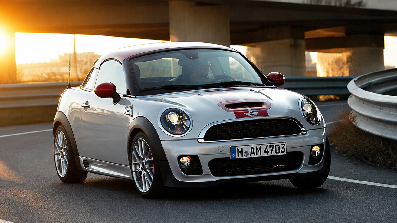 Carro esportivo Mini Cooper prata com faixa vermelha dirigindo em estrada curva ao pôr do sol.