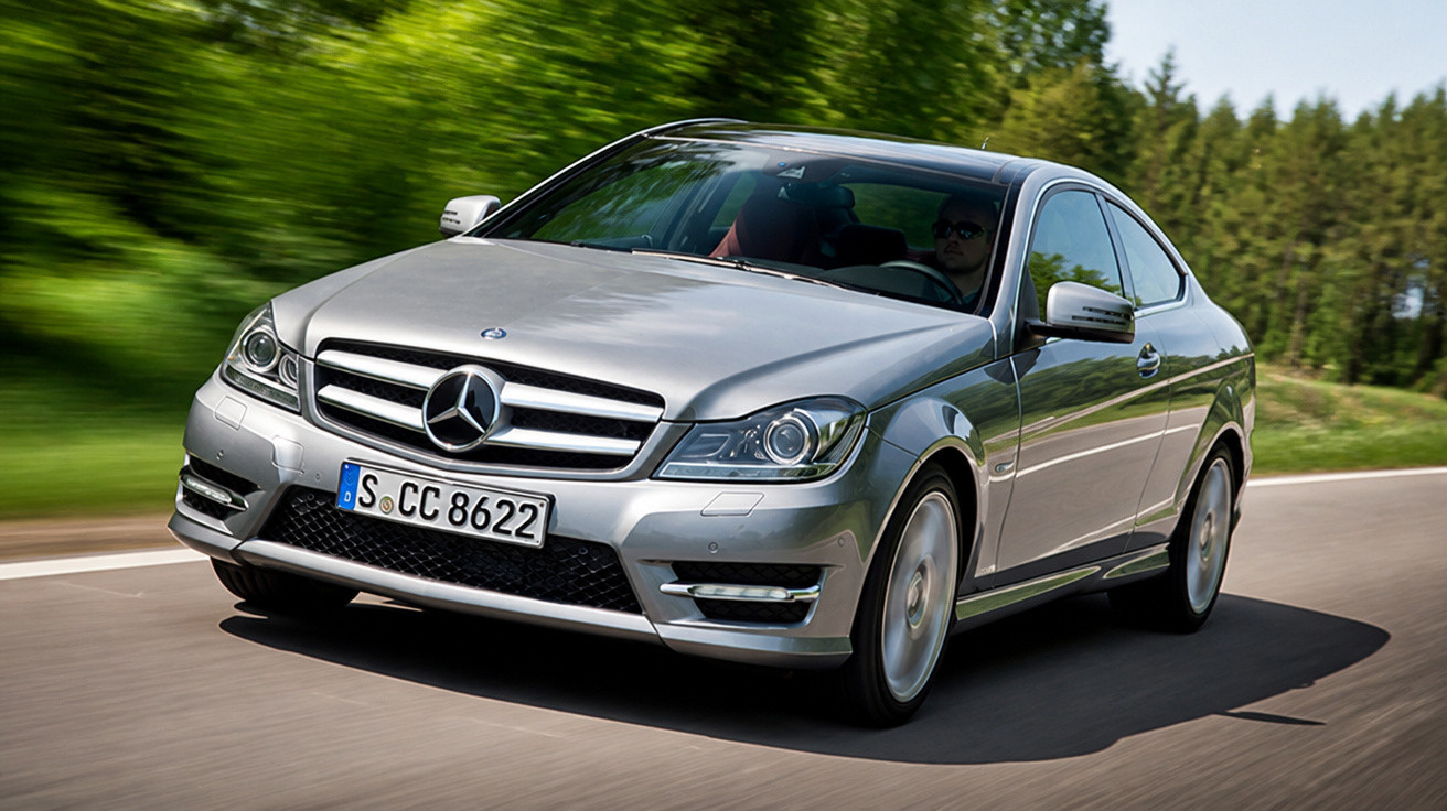 Carro prata Mercedes-Benz em movimento em estrada rodeada por árvores verdes em dia ensolarado.