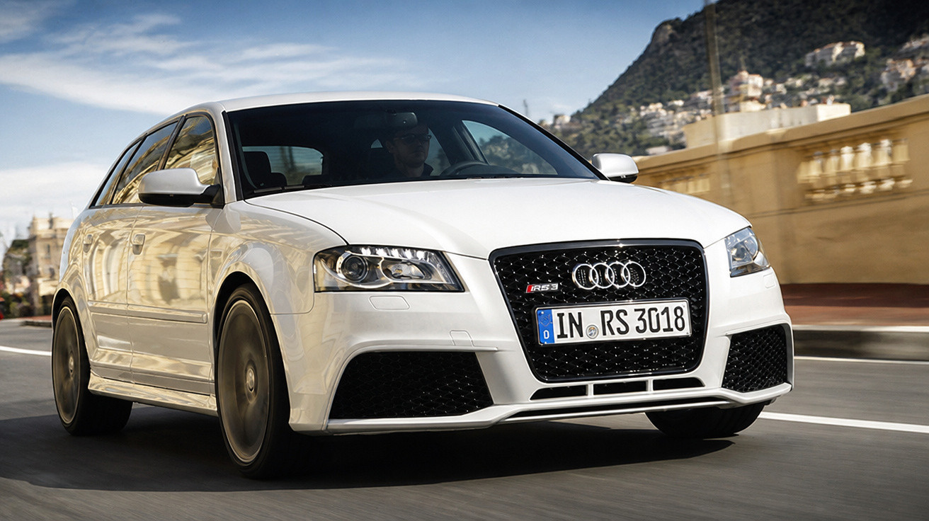 Carro esportivo Audi RS branco em movimento em estrada urbana com edifícios ao fundo.