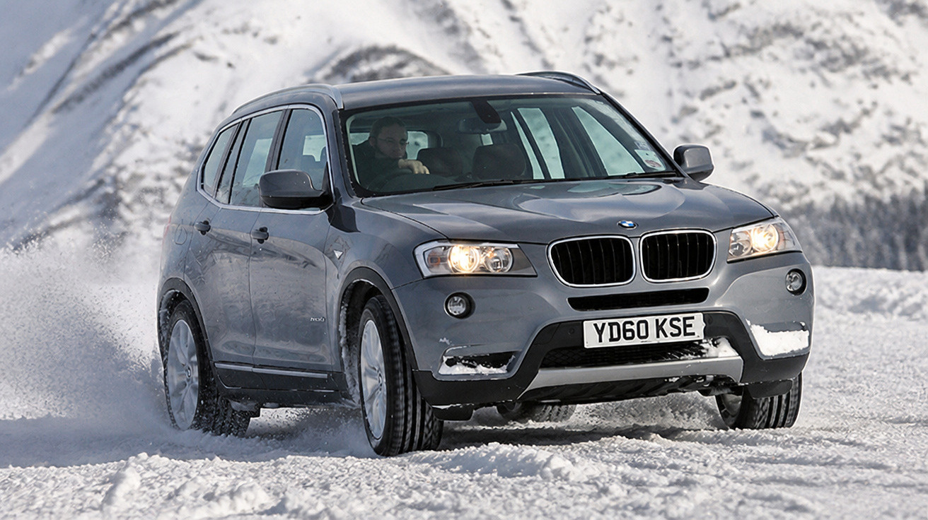 Carro SUV cinza dirigindo na neve com montanhas ao fundo em dia claro.