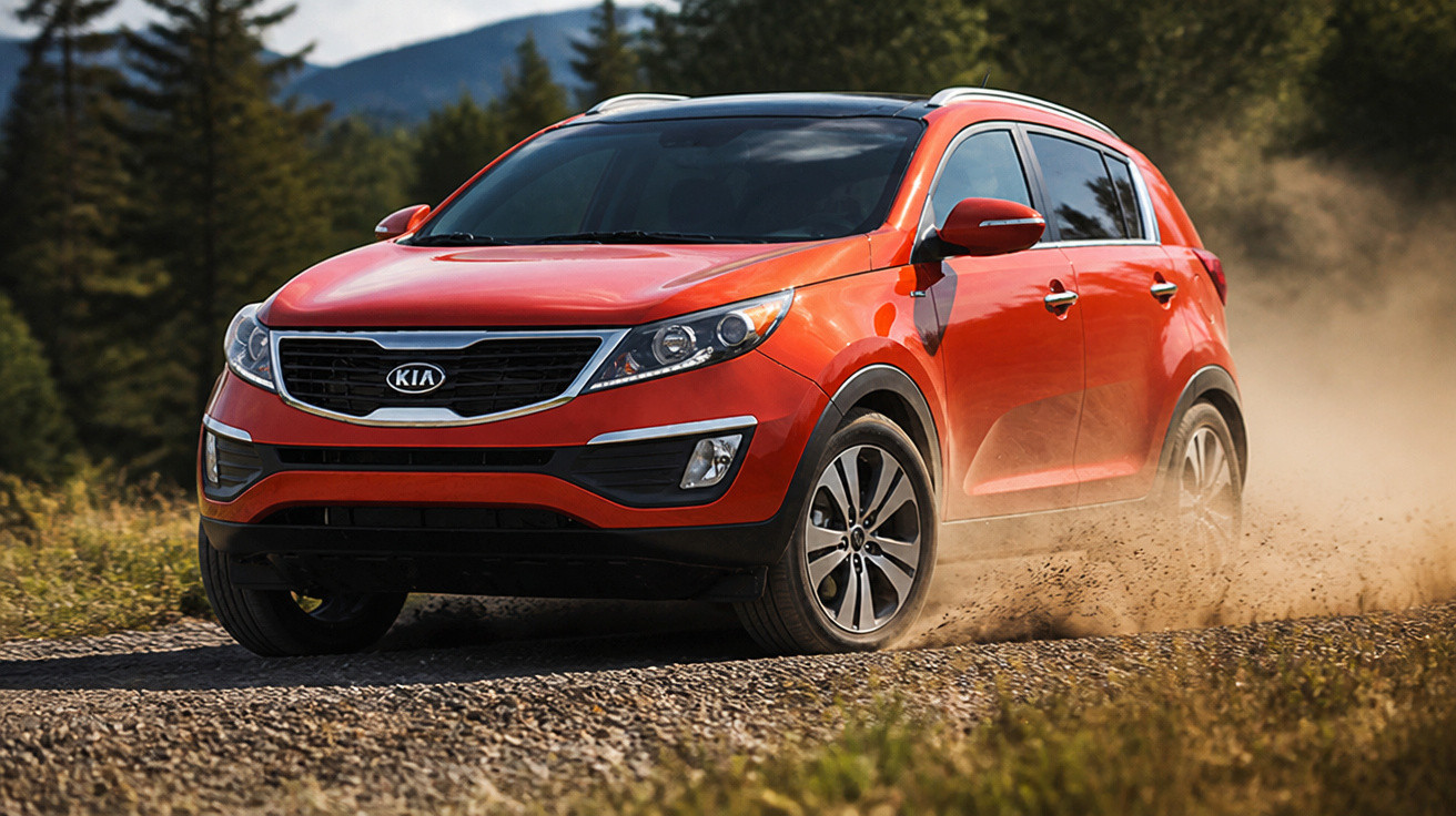 Carro SUV vermelho Kia em estrada de terra com poeira e árvores ao fundo em dia ensolarado.