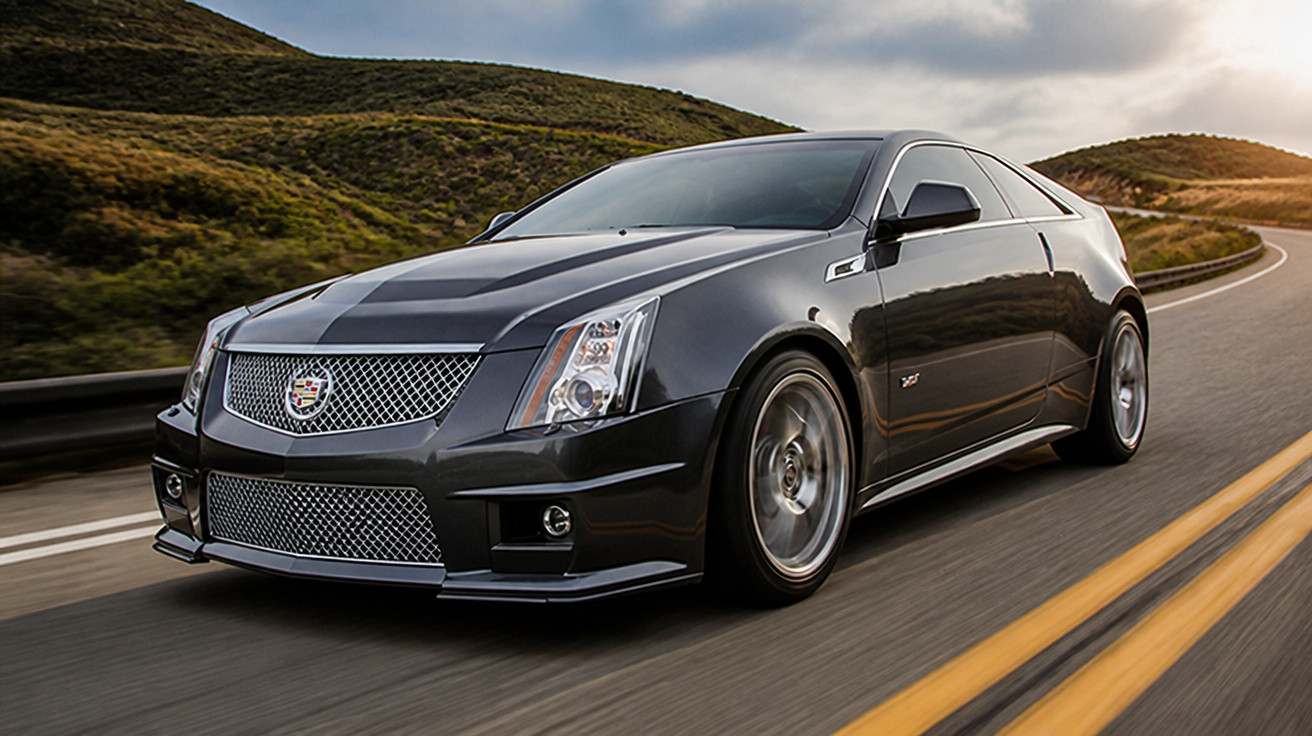 Carro esportivo preto Cadillac em estrada sinuosa com colinas verdes ao fundo, em movimento ao pôr do sol.