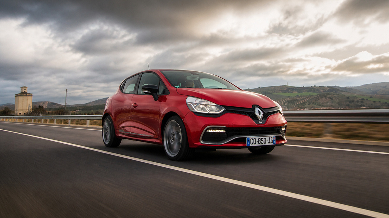 Carro hatch vermelho Renault trafegando em estrada com céu nublado e colinas ao fundo.