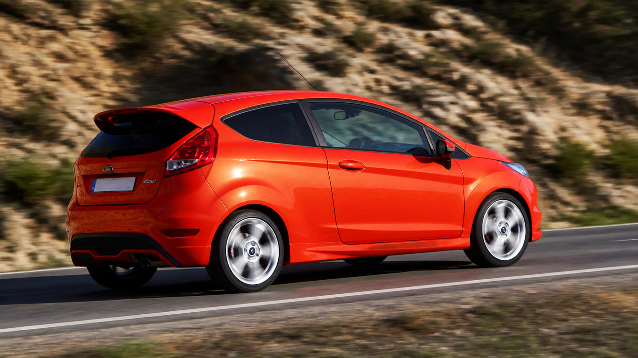 Carro hatchback vermelho em movimento em estrada asfaltada próximo a montanha rochosa.