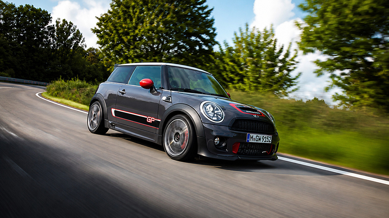 Carro esportivo Mini Cooper GP cinza com detalhes vermelhos dirigindo em estrada curva cercada por árvores.