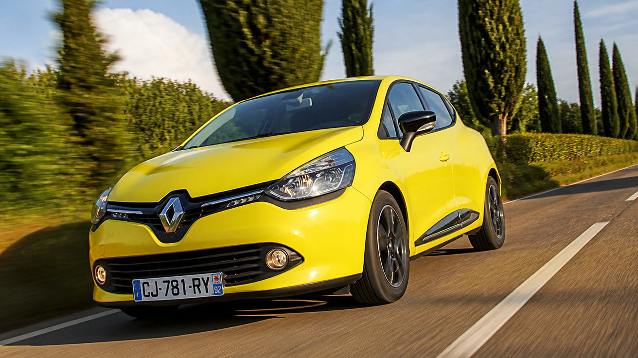 Carro amarelo Renault Clio em estrada reta cercada por árvores altas e céu azul.