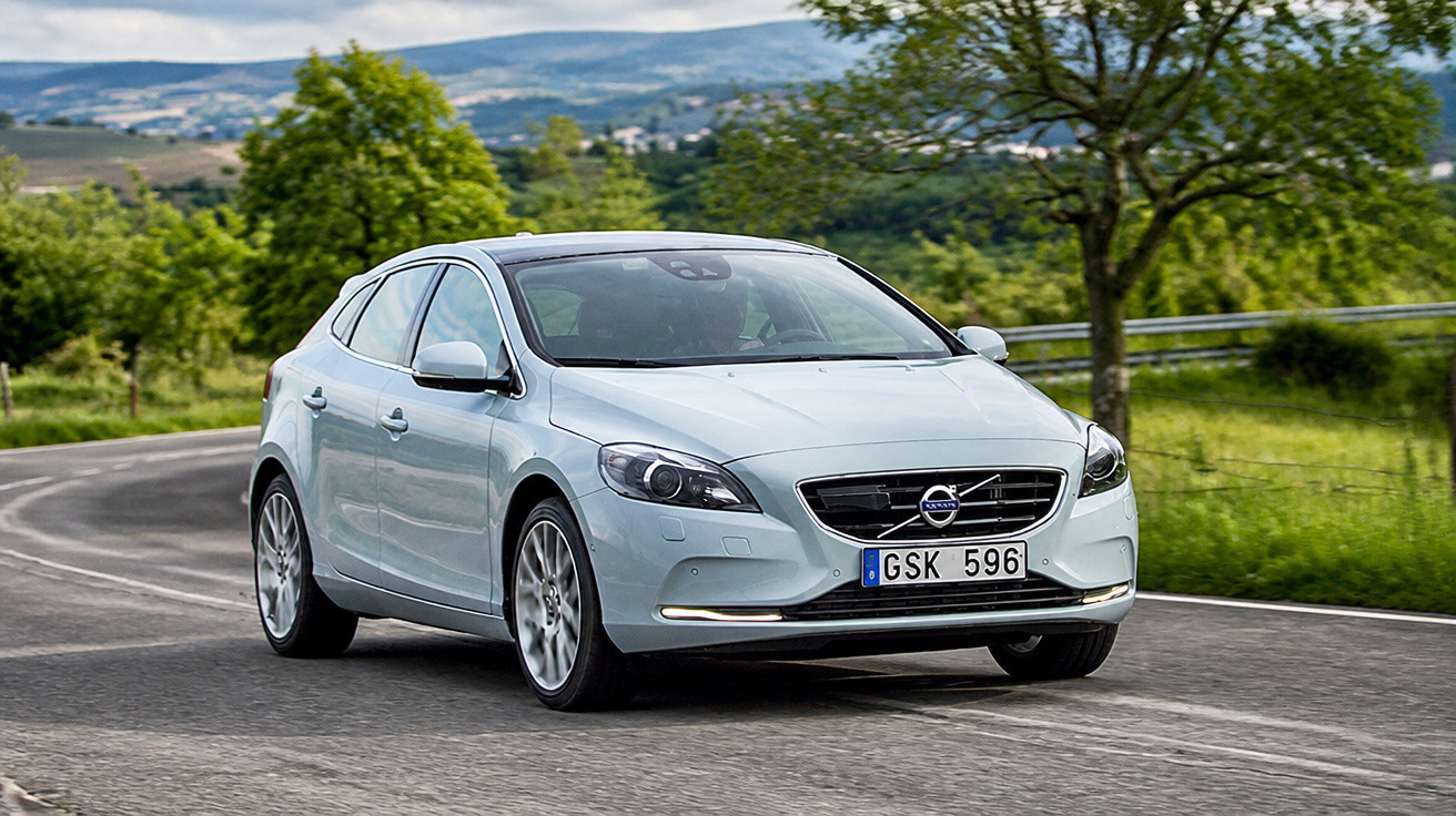 Carro prata modelo Volvo em curva de estrada asfaltada com árvores e morros ao fundo.