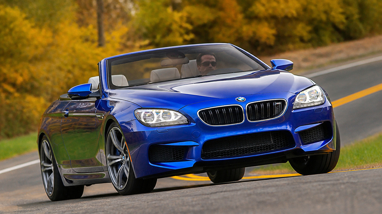 Carro conversível azul da BMW em movimento em estrada com árvores amarelas ao fundo.