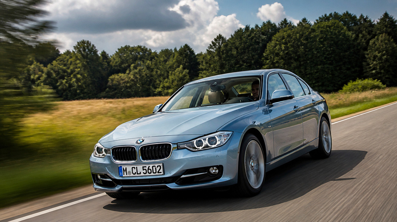 Carro BMW cinza em movimento numa estrada cercada por árvores sob céu parcialmente nublado.