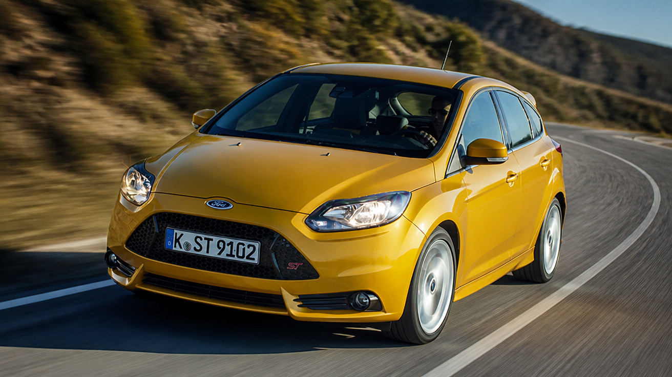 Carro hatchback amarelo Ford Focus ST em movimento em estrada sinuosa com colinas ao fundo.
