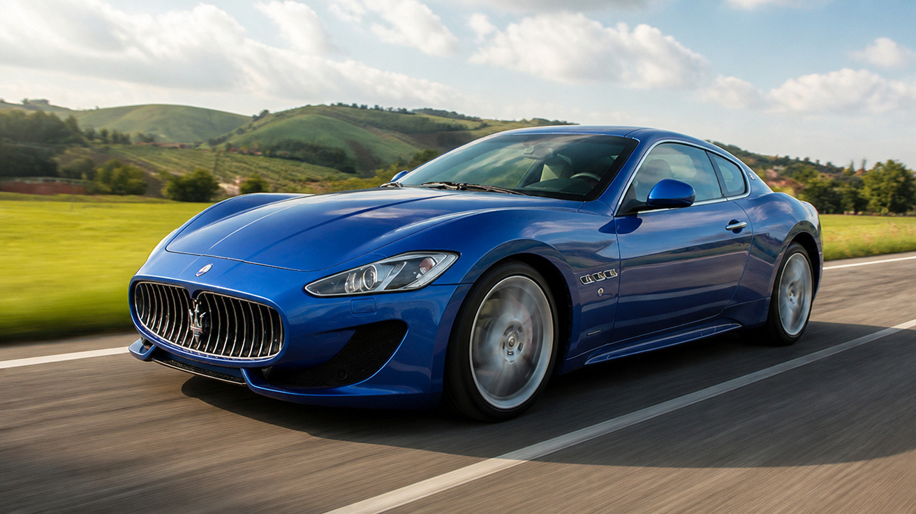 Carro esportivo azul Maserati em movimento em estrada com colinas verdes ao fundo sob céu azul com nuvens.