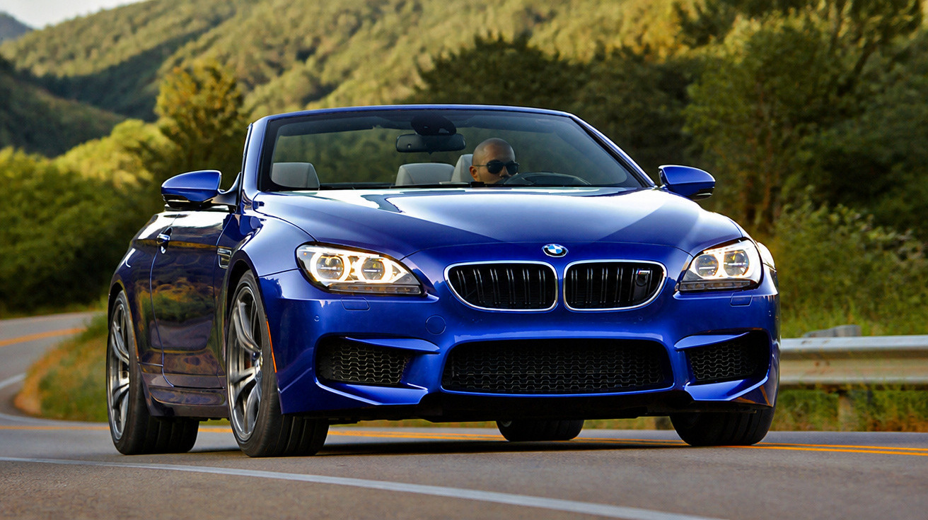 Carro esportivo conversível azul BMW com motorista de óculos em estrada sinuosa cercada por árvores.