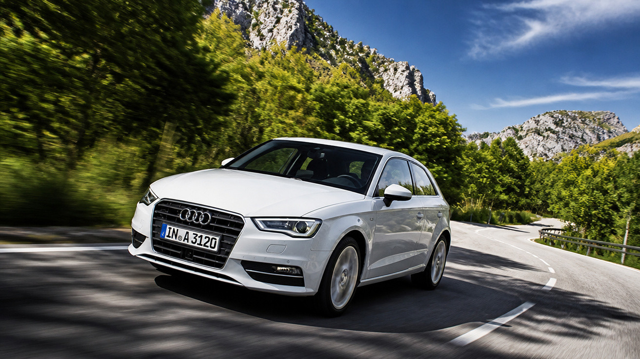 Carro Audi branco em alta velocidade em estrada sinuosa cercada por árvores e montanhas.
