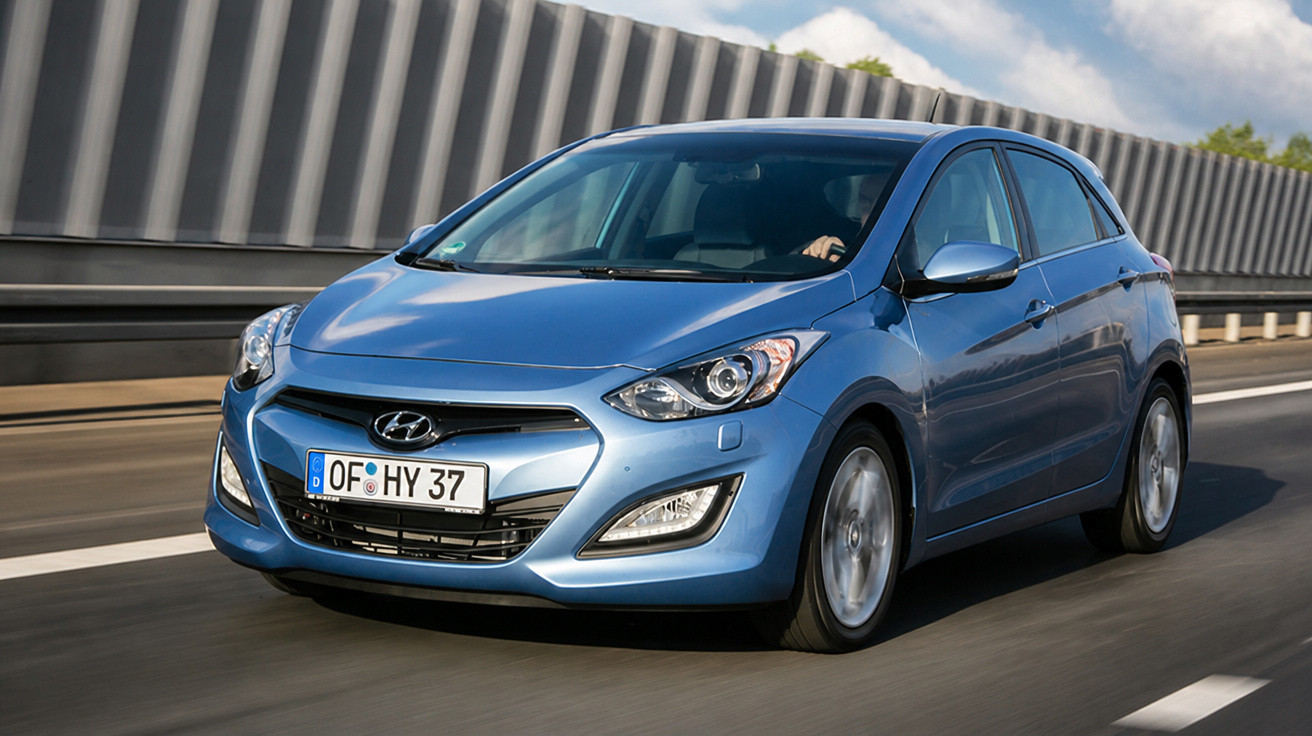 Carro hatchback azul Hyundai em movimento na estrada com barreira de concreto ao fundo.