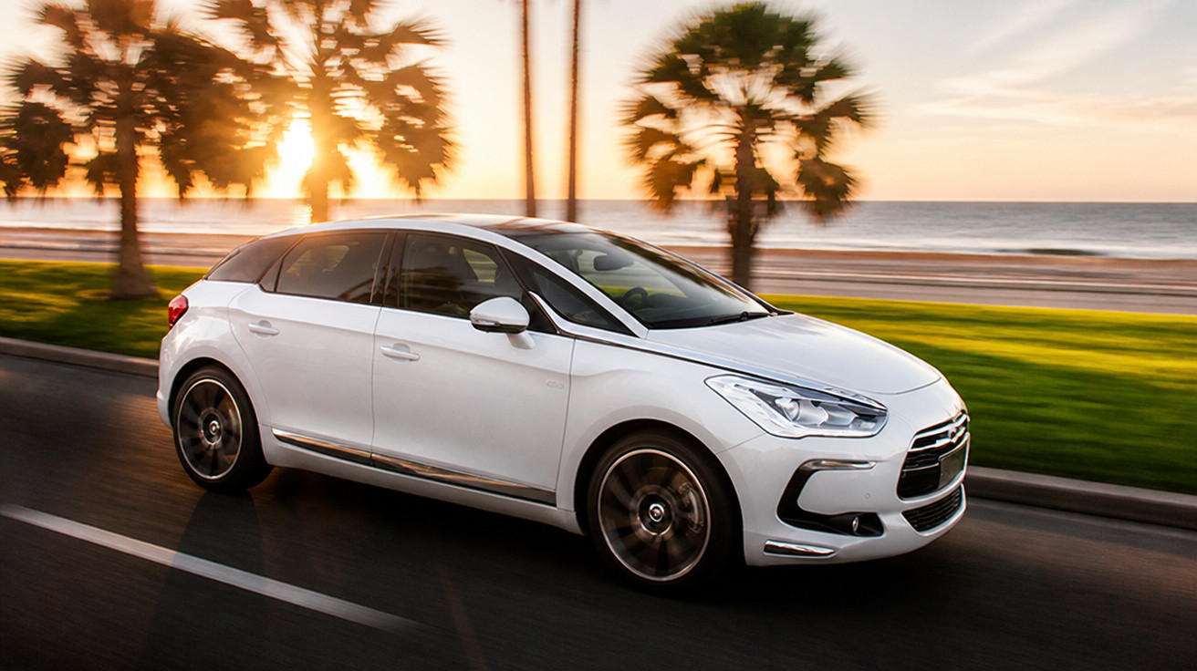Carro branco em movimento à beira-mar ao pôr do sol com palmeiras ao fundo.