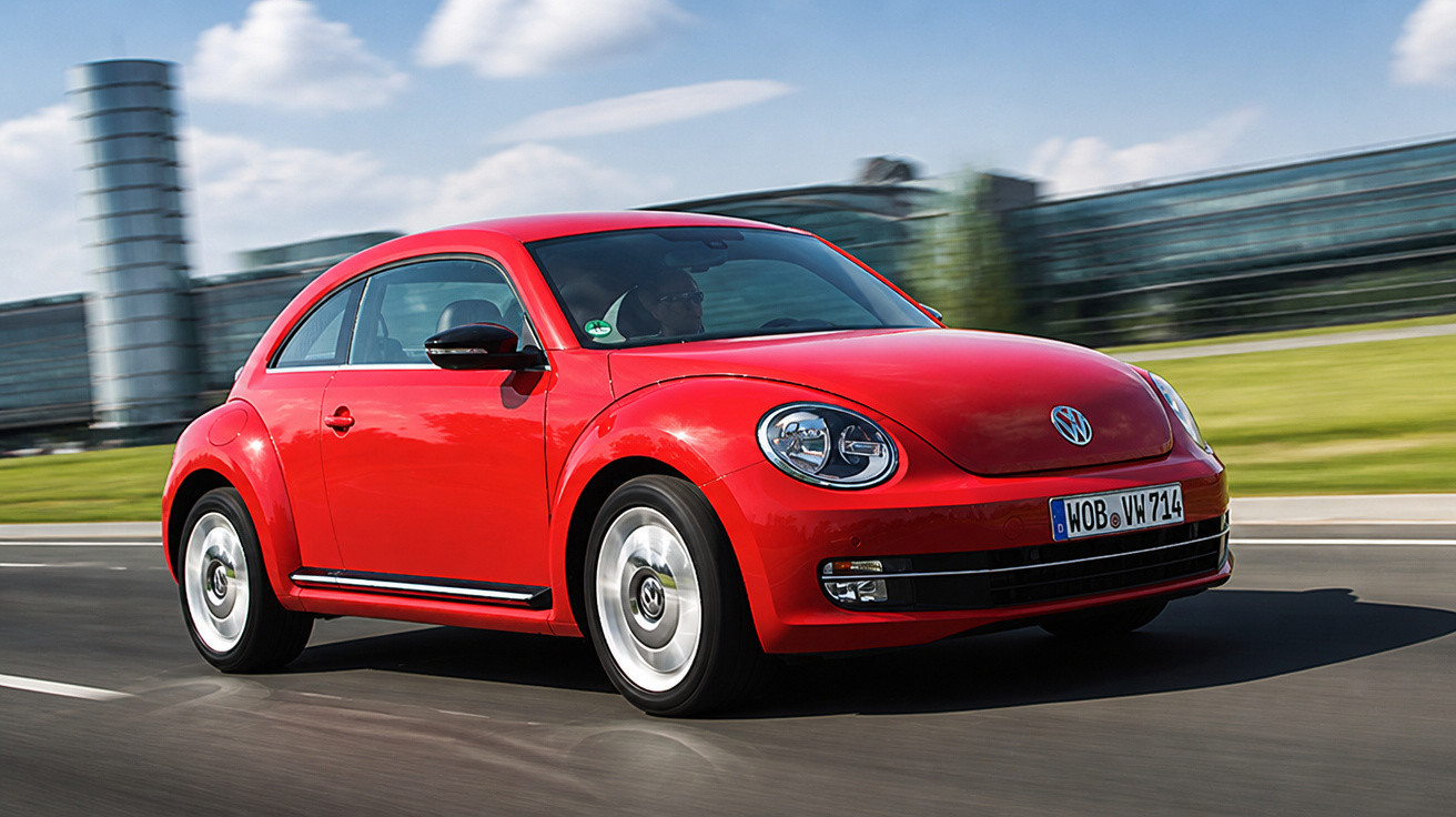 Carro Volkswagen Fusca vermelho em movimento em uma estrada urbana com prédios ao fundo.