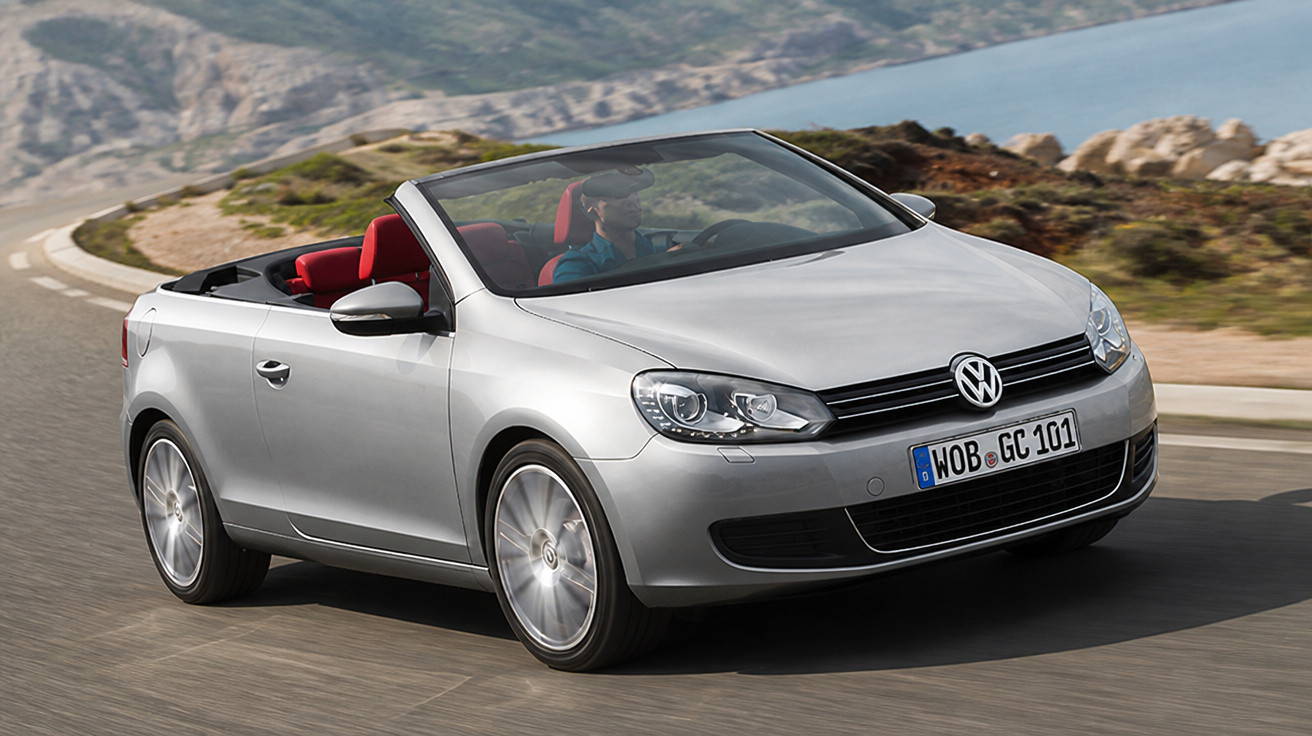 Carro conversível prata Volkswagen com interior vermelho dirigindo em estrada sinuosa com paisagem montanhosa ao fundo.