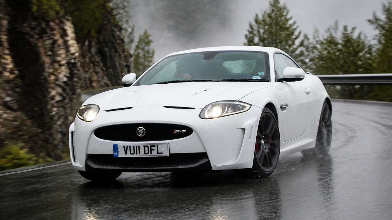 Carro esportivo branco Jaguar dirigindo em estrada molhada com neblina e árvores ao fundo.