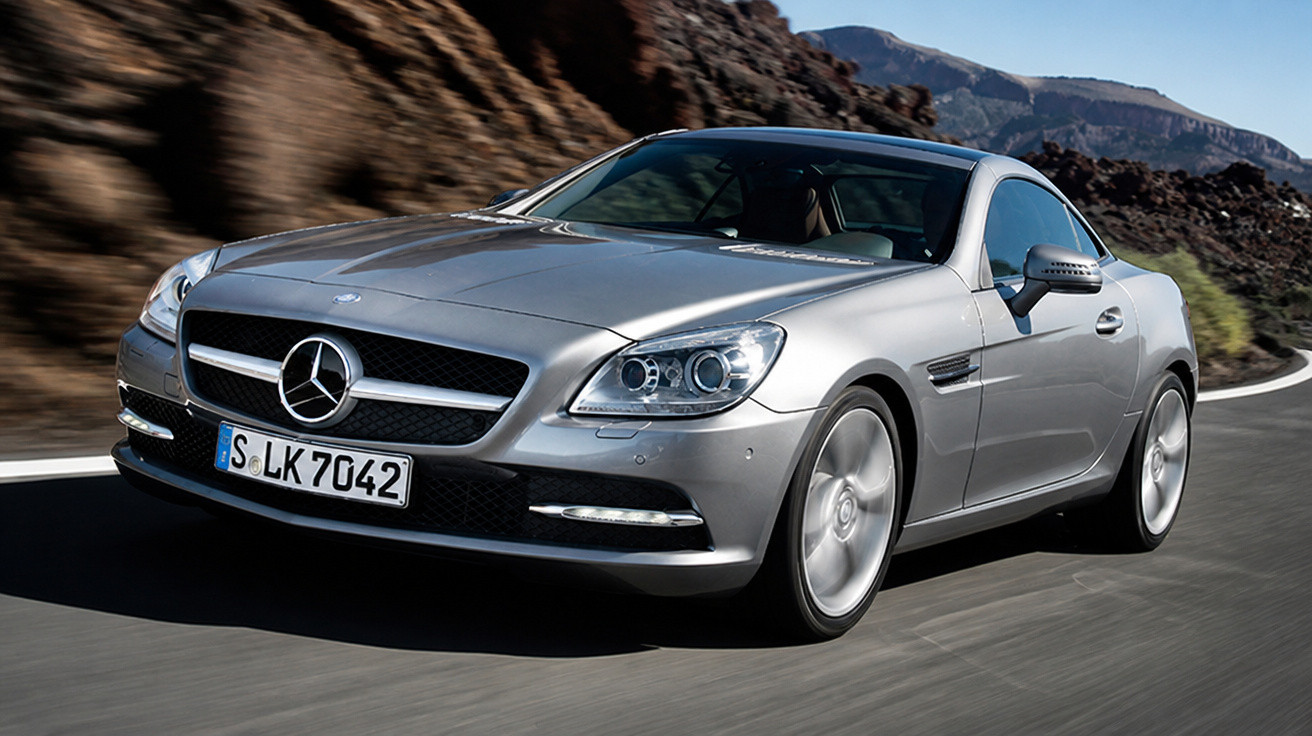 Carro esportivo prata Mercedes-Benz em estrada sinuosa com montanhas ao fundo em dia ensolarado.