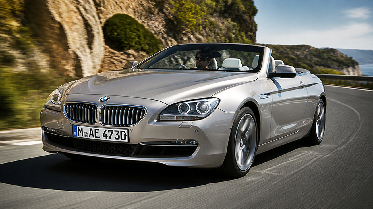 Carro conversível BMW prata dirigido em estrada sinuosa com paisagem montanhosa ao fundo.