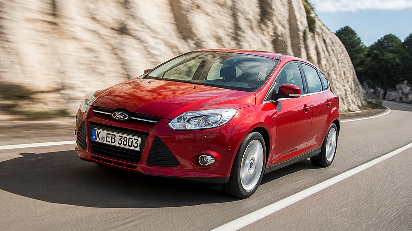 Carro hatchback vermelho Ford em movimento em estrada sinuosa com paredes rochosas ao fundo.