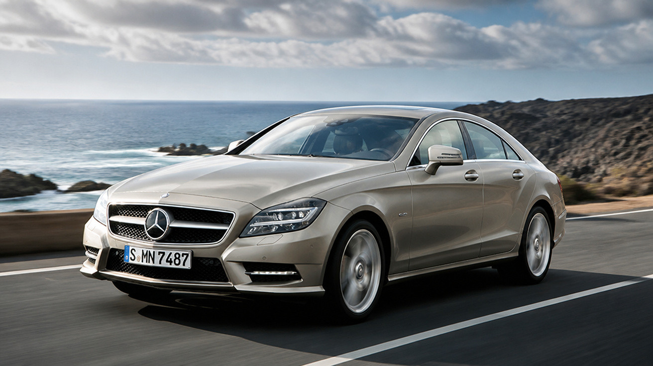 Carro sedan prata Mercedes-Benz em estrada costeira com mar e céu nublado ao fundo.