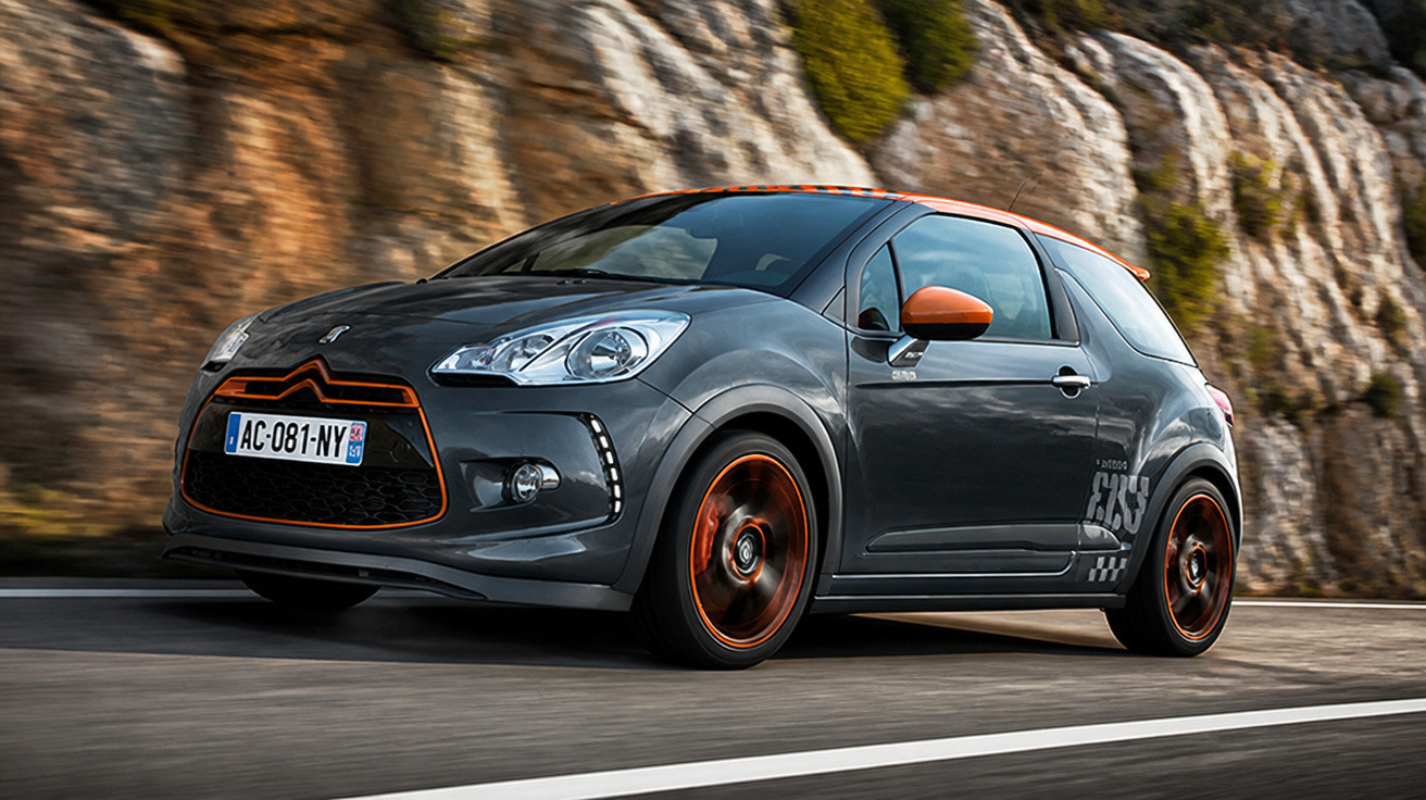 Carro Citroën DS3 cinza com detalhes laranja trafegando em estrada ao lado de encosta rochosa.
