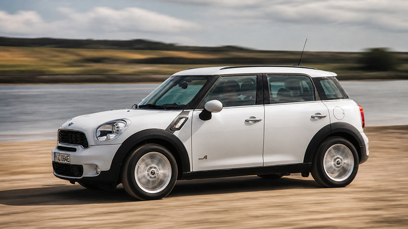 Carro branco MINI Countryman em movimento na beira de um lago com céu nublado ao fundo.