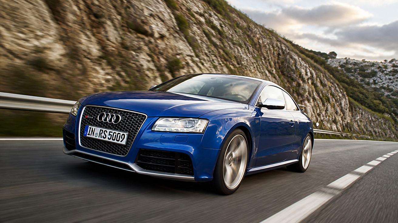 Carro esportivo Audi azul acelerando em estrada com montanhas rochosas ao fundo.