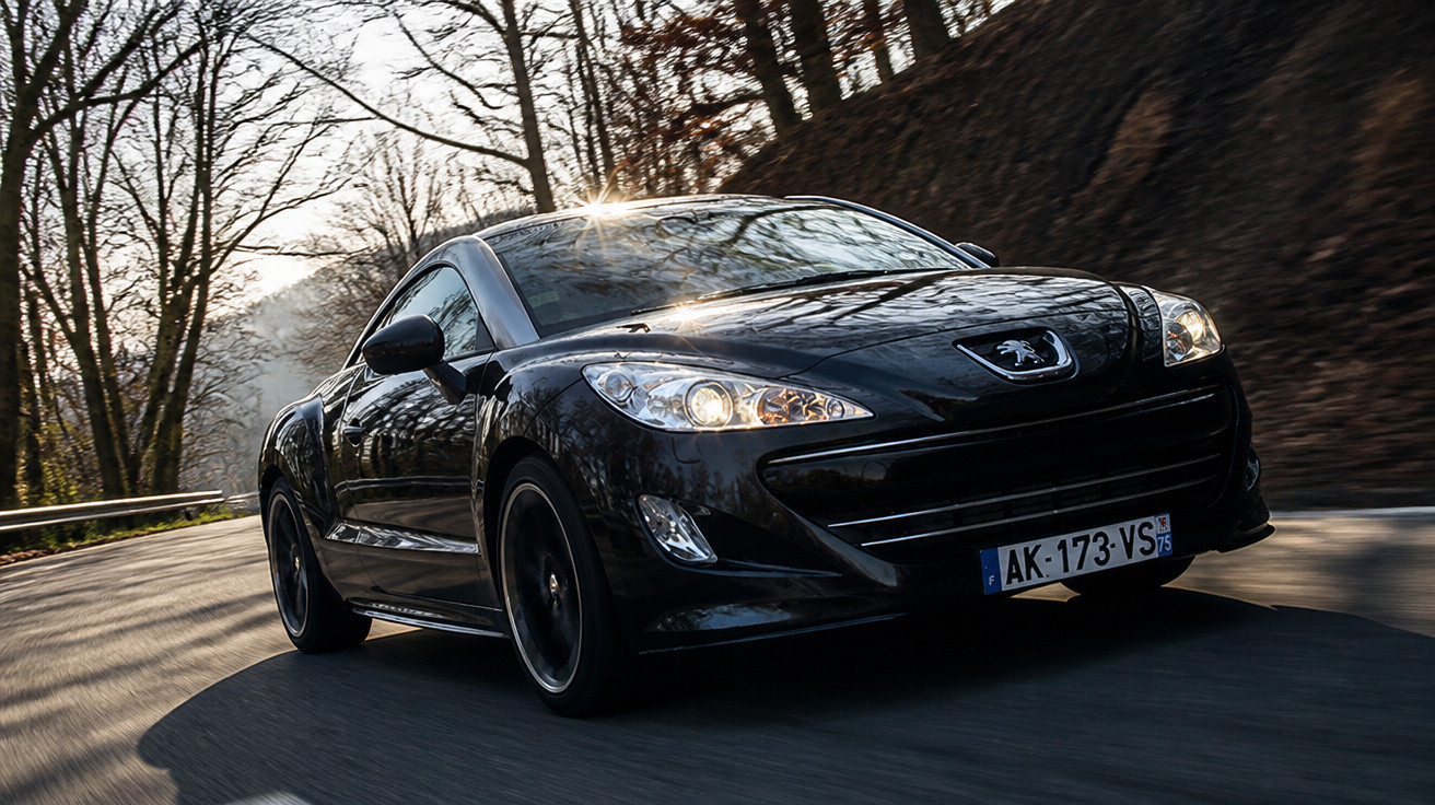 Carro esportivo preto Peugeot circulando em estrada sinuosa cercada por árvores sem folhas ao amanhecer.