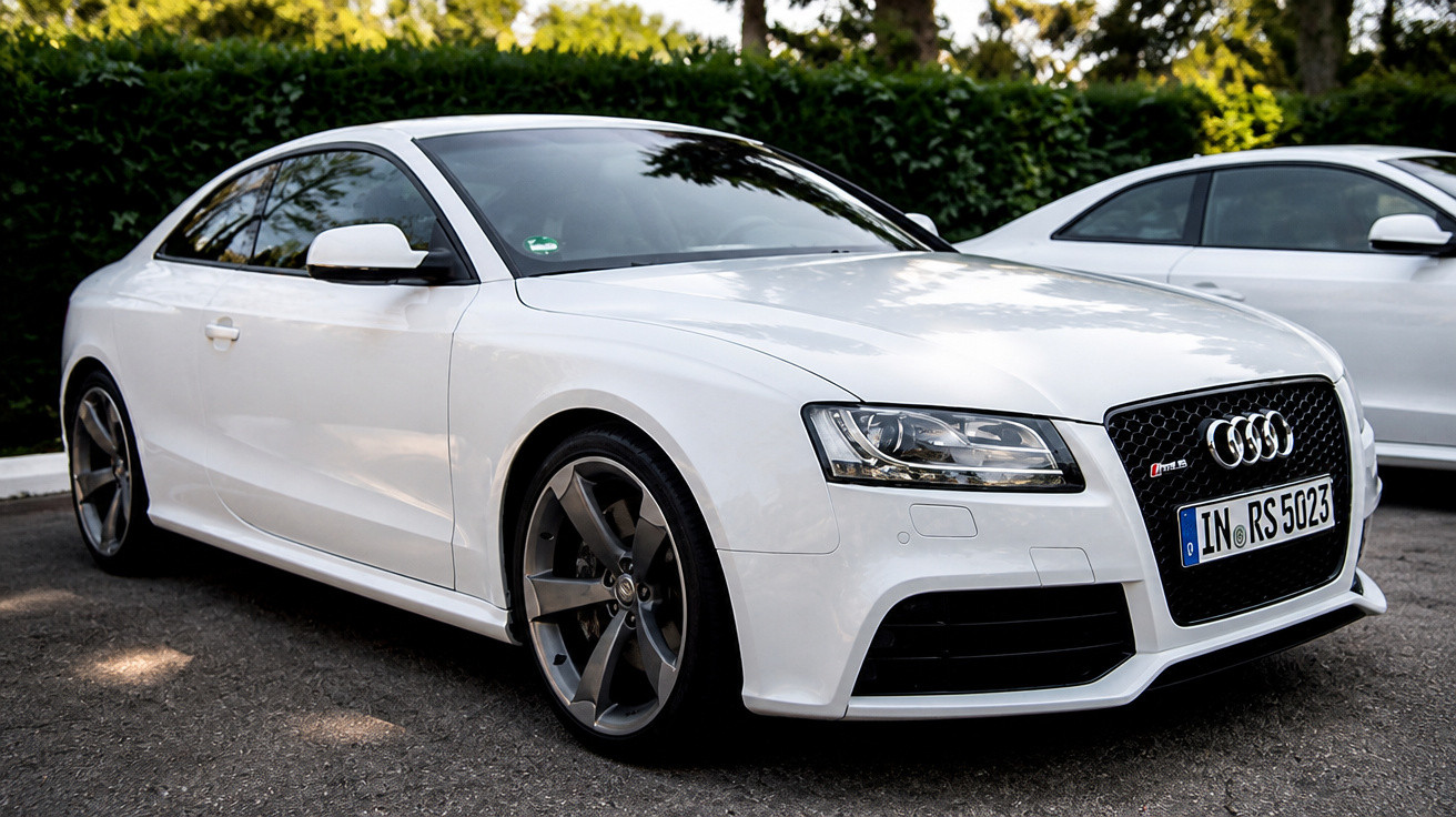 Carro esportivo branco Audi RS5 estacionado em área externa com vegetação ao fundo.