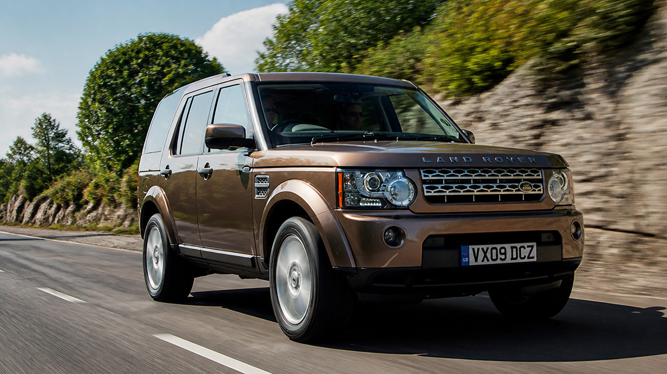 Carro SUV Land Rover marrom em movimento em estrada com árvores e muro de pedra ao fundo.