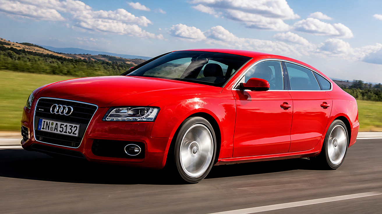 Carro vermelho Audi sedan em movimento em estrada com céu azul e campo ao fundo.