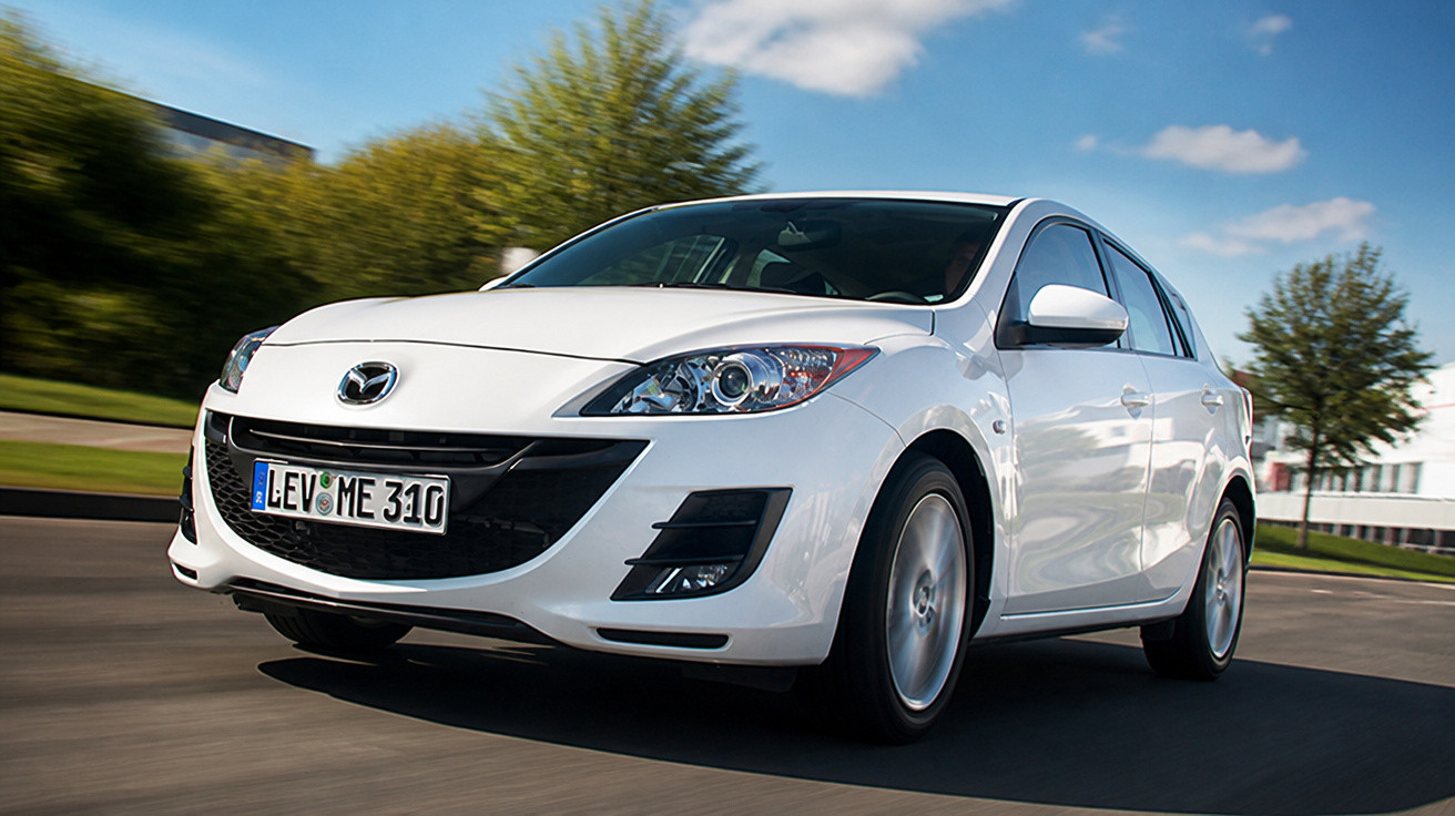 Carro branco Mazda em movimento em estrada com árvores e céu azul ao fundo.