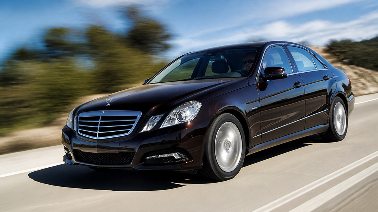 Carro preto Mercedes-Benz sedã em movimento em estrada com céu azul ao fundo.