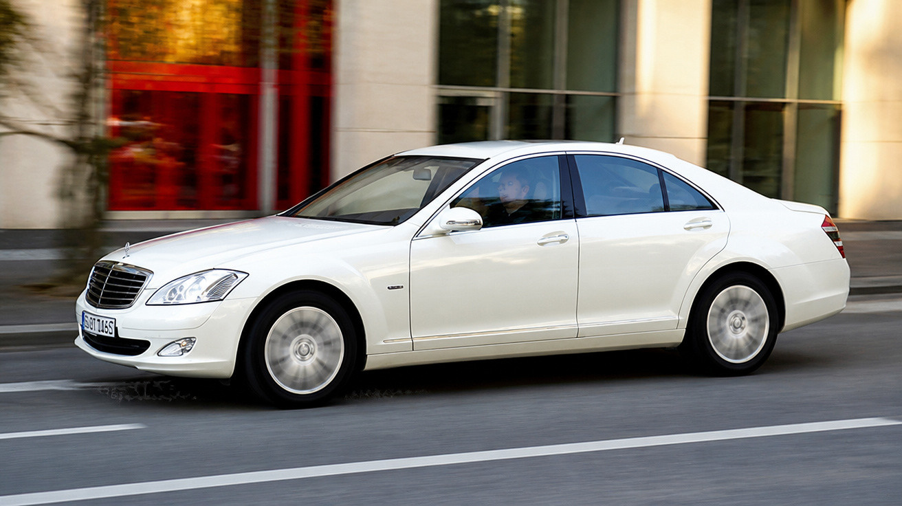 Carro sedã branco da Mercedes-Benz em movimento na rua com prédios ao fundo.