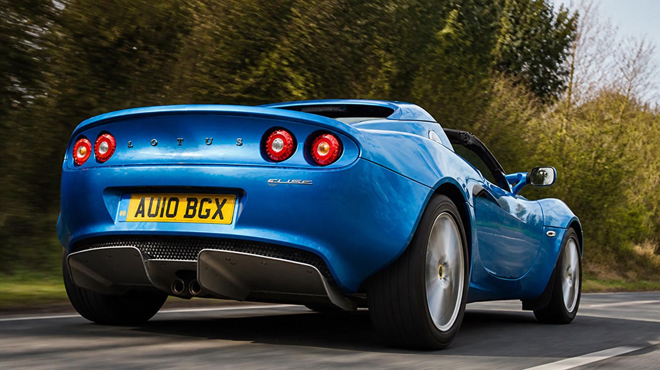 Carro esportivo azul modelo Lotus Elise em movimento em estrada cercada por árvores.