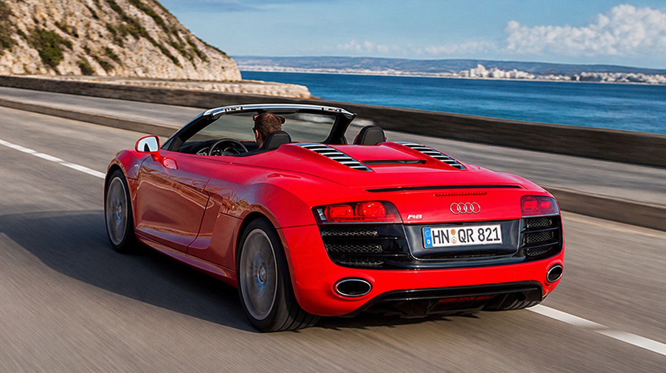 Carro esportivo conversível vermelho Audi R8 dirigindo em estrada à beira-mar em dia ensolarado.