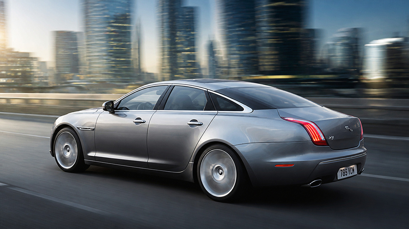 Carro cinza prata sedan de luxo em movimento com edifícios modernos desfocados ao fundo.